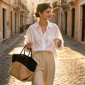 May include: A white long-sleeved button-down shirt with a chest pocket, beige pleated trousers, and a large two-toned tote bag featuring a black leather upper section and a woven tan straw base, held by a person in a sunlit cobblestone street.