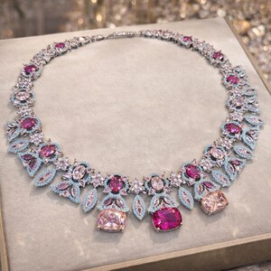 Puede incluir: Un collar plateado con un dise&ntilde;o floral, con piedras preciosas rosas y celeste. El collar tiene una serie de piedras rosas y transparentes, con piedras rectangulares rosas y melocot&oacute;n m&aacute;s grandes como puntos focales. El collar se exhibe sobre una superficie neutra.