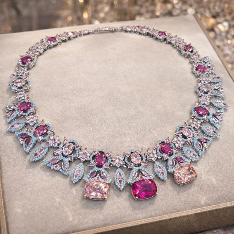 Puede incluir: Un collar plateado con un dise&ntilde;o floral, con piedras preciosas rosas y celeste. El collar tiene una serie de piedras rosas y transparentes, con piedras rectangulares rosas y melocot&oacute;n m&aacute;s grandes como puntos focales. El collar se exhibe sobre una superficie neutra.