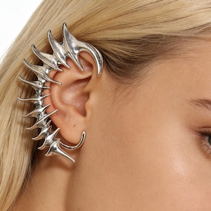 May include: Silver ear cuff with a spiky, organic design. The ear cuff curves around the ear and features a series of pointed, thorn-like elements. The jewelry is worn on the ear and is a unique accessory.