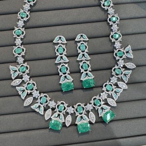 Puede incluir: Un conjunto de collar y pendientes plateados. El collar y los pendientes presentan piedras preciosas de color verde esmeralda y detalles claros y brillantes. El collar tiene un dise&ntilde;o floral con esmeraldas m&aacute;s grandes en el centro.