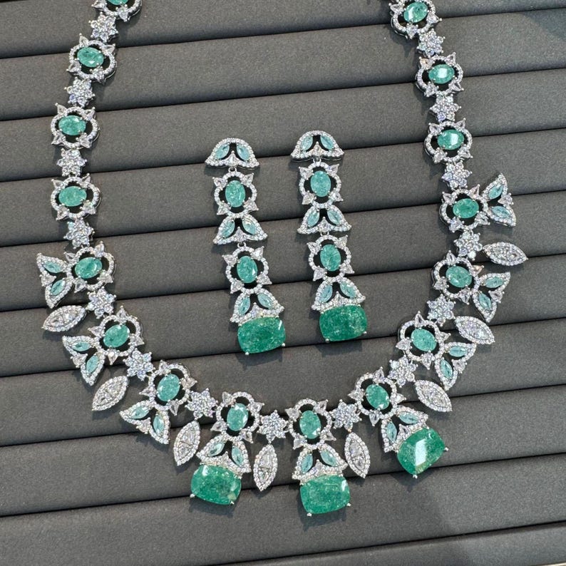 Puede incluir: Un conjunto de collar y pendientes plateados. El collar y los pendientes presentan piedras preciosas de color verde esmeralda y detalles claros y brillantes. El collar tiene un dise&ntilde;o floral con esmeraldas m&aacute;s grandes en el centro.
