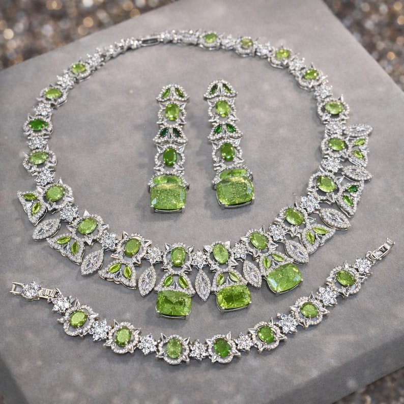Puede incluir: Un conjunto de joyas plateadas que incluye collar, pendientes y pulsera. El conjunto est&aacute; adornado con piedras preciosas verdes y cristales transparentes. El collar y la pulsera tienen un dise&ntilde;o floral.