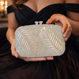 May include: A rectangular clutch bag featuring a cream-coloured satin base adorned with intricate silver-toned leaf patterns and dense, iridescent crystal embellishments. The accessory includes a metallic clasp closure at the top centre.