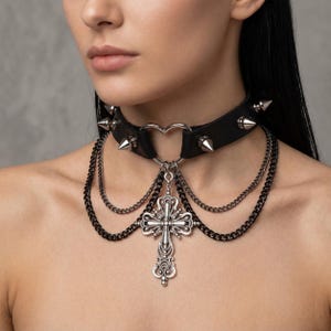 May include: Black leather choker necklace with silver-toned spikes and a heart-shaped center. The necklace features layered chains and a decorative cross pendant, creating a gothic or alternative aesthetic.