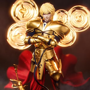 Op de afbeelding: Een gouden beeldje van een personage met blond haar en rode ogen, gekleed in een sierlijk gouden harnas. De figuur heeft de armen gekruist en staat tegen een achtergrond van gloeiende gouden bollen en een rode cape.