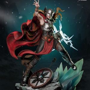 May include: A detailed figurine of a female warrior in a dynamic pose, clad in armor and a red cape. She holds a hammer and is standing on a rocky base with a shield and arrows. The figure has a helmet and flowing blonde hair.