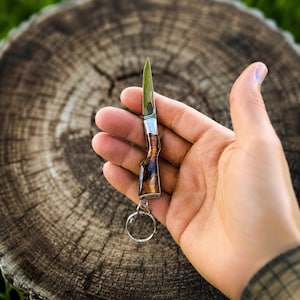 May include: A miniature folding pocket knife keyring featuring a handle shaped like a rifle stock with a polished wood grain finish and a silver metal bolster. The small steel blade is extended, and a circular metal ring is attached to the base.