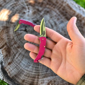 Puede incluir: Una pequeña navaja plegable con mango de color rosa brillante y hoja de metal plateado. La hoja presenta un pequeño logotipo grabado. Una segunda navaja plegable rosa idéntica es visible al fondo. El mango está asegurado con dos remaches de latón.