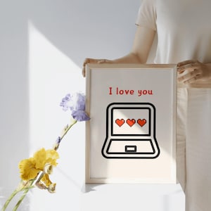 May include: A framed print with the words "I love you" in red above a laptop graphic with three red hearts on the screen. The print is held by a person. A vase of flowers is in the foreground.