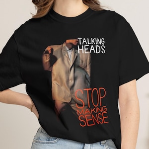 May include: A black short-sleeved t-shirt featuring a graphic print of a person wearing an oversized beige suit. The text TALKING HEADS appears in white above the figure, and the words STOP MAKING SENSE are printed in red below the figure.