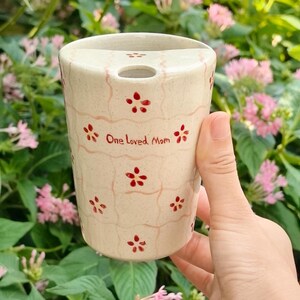 May include: A cream-colored ceramic travel mug with a speckled finish, featuring a light pink grid pattern and small red flower motifs. The text "One Loved Mom" is printed in red on the front, and the mug has a lid with a drinking hole.