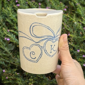May include: A cylindrical ceramic cup with a speckled off-white finish and a flat lid. The exterior features a hand-drawn blue ink design of two connected heart-shaped lockets tied with a ribbon, with the letters C and K inside the hearts.