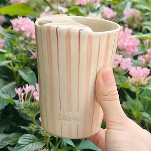 Peut inclure: Un gobelet de voyage cylindrique en céramique avec un émail crème moucheté et des rayures verticales roses. Le gobelet présente un couvercle moulé avec une ouverture pour boire et le texte "Julia's Mom" inscrit près de la base.