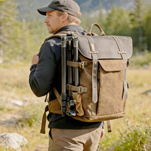 Peut inclure: Un sac à dos marron et noir avec un trépied attaché. Le sac à dos a des sangles en cuir et une fermeture à rabat. La personne porte une casquette et une veste noires, et un pantalon kaki. L'arrière-plan est une scène extérieure floue.