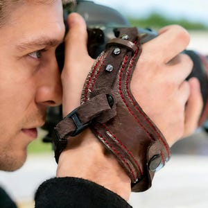 May include: Brown leather camera wrist strap with red stitching and silver rivets. The strap has a black buckle and is designed to secure a camera to the user's wrist. The strap is shown being used with a DSLR camera.