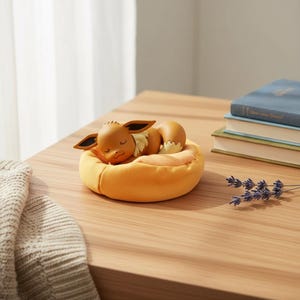 Op de afbeelding: Een slapende Eevee-figuur rust in een geel bed op een houten tafel. Het beeldje is bruin met grote oren en een donzige crèmekleurige nek. Een stapel boeken en een takje lavendel liggen ook op de tafel. Het bed heeft een diameter van ongeveer 10 cm.