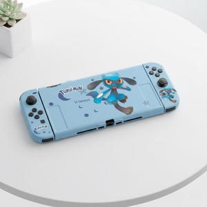 Puede incluir: Una consola Nintendo Switch azul claro con un personaje de dibujos animados de Pokémon azul y negro. La consola tiene botones negros y un pequeño logotipo de Pokémon.
