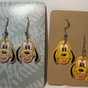 May include: Two pairs of dangle earrings featuring a cartoon dog face design. One pair is light tan with black accents and pink tongues, the other is yellow with black accents and pink tongues. Both pairs have silver-coloured ear wires.