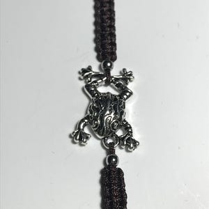 Peut inclure: Un charm argenté en forme de grenouille est suspendu à un cordon tressé marron foncé, avec un pompon assorti. Le charm de la grenouille présente des détails et est maintenu entre deux petites perles argentées.