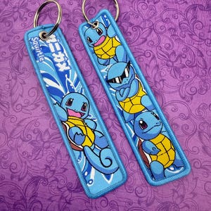 May include: Two blue keychains featuring the character Squirtle from the Pokémon franchise. Each keychain has a blue background with a Squirtle design and a silver keyring. One keychain has the word "Squirtle" and Japanese text.