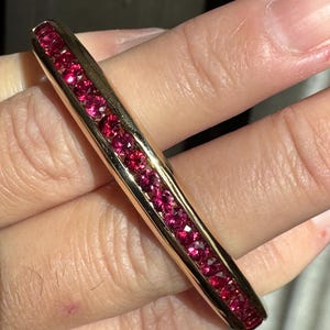 Pode incluir: Uma pulseira dourada com uma fileira de pedras preciosas vermelhas vibrantes. As pedras preciosas são colocadas próximas umas das outras, criando uma linha contínua de cor. A pulseira tem um acabamento polido, refletindo a luz.