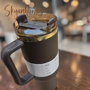 May include: A dark brown insulated tumbler with a sturdy handle and a transparent amber-tinted lid featuring a black sliding closure. A white paper band with printed text and icons wraps around the lower section of the matte-finished vessel.