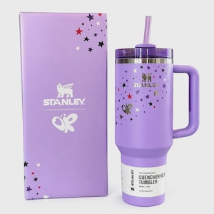 Puede incluir: Un vaso Stanley Quencher H2.0 de color morado claro con una capacidad de 1,18 litros, que incluye pajita, asa a juego y un dise&ntilde;o de estrellas. El vaso se muestra junto a su caja morada clara a juego, que tambi&eacute;n presenta gr&aacute;ficos de estrellas.