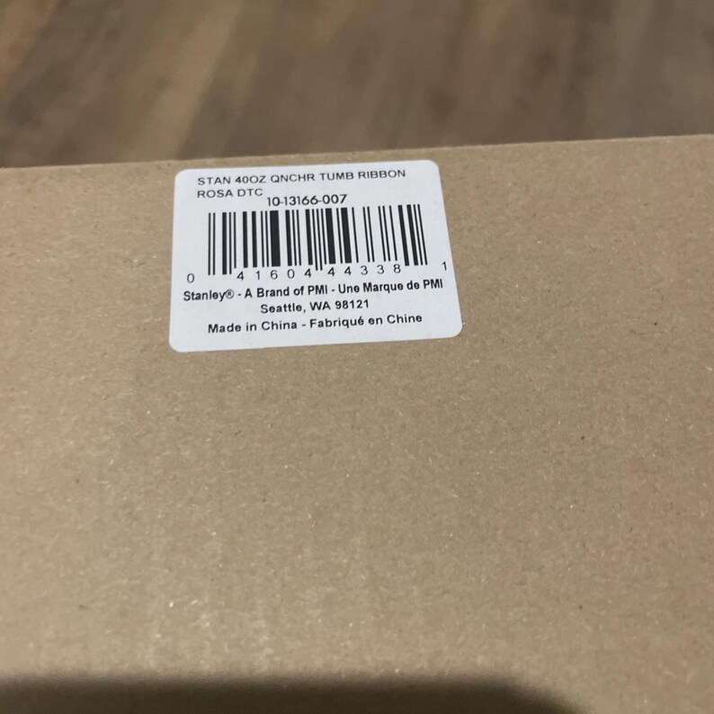 May include: A tan cardboard box featuring a white rectangular product label with a barcode and the text: STAN 40OZ QNCHR TUMB RIBBON ROSA DTC, 10-13166-007, Stanley - A Brand of PMI, Seattle, WA 98121, Made in China - Fabriqu&eacute; en Chine.