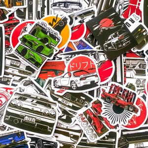 Puede incluir: Una colección de pegatinas con temática de coches que presentan varios modelos en negro, blanco, rojo y verde. Algunas pegatinas incluyen texto japonés e imágenes del sol. Las pegatinas son de diferentes formas y tamaños.