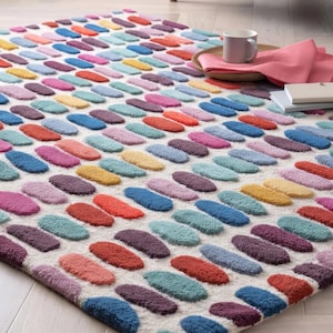 May include: A rectangular area rug with a repeating pattern of colorful, oval-shaped tufts. The rug features a cream-colored background with tufts in shades of red, orange, blue, purple, pink, and yellow. The rug is placed on a light-colored wooden floor.