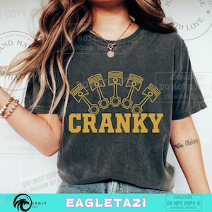 May include: Dark gray t-shirt featuring a gold graphic of engine pistons above the word "CRANKY" in gold. The shirt is a casual style, suitable for everyday wear. The design is printed on the front of the shirt.