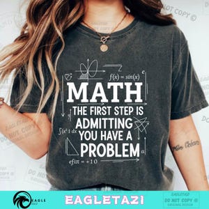 May include: Dark grey t-shirt featuring the word "MATH" in white, accompanied by mathematical symbols and the statement "THE FIRST STEP IS ADMITTING YOU HAVE A PROBLEM." A casual, comfortable style.