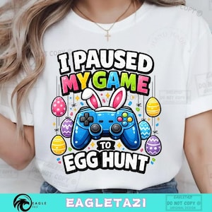 Könnte beinhalten: Weißes T-Shirt mit einer bunten Grafik, die einen blauen Videospiel-Controller mit Hasenohren zeigt, umgeben von Ostereiern. Der Text lautet "I PAUSED MY GAME TO EGG HUNT". Das Design ist lebendig und verspielt, ideal für Ostern.