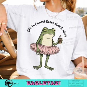 May include: White t-shirt featuring a green frog in a pink tutu, holding a coffee cup. Text above the frog reads "Off to Commit Dance Mom Foolery." The shirt has a relaxed fit and a crew neck.
