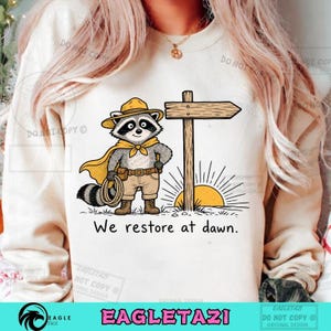 Puede incluir: Una sudadera color crema con el gráfico de un mapache con sombrero amarillo, capa y pantalones cortos color canela, junto a un poste de madera y un sol naciente. El texto "We restore at dawn." está impreso en negro bajo la ilustración.