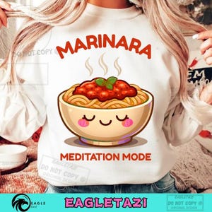 May include: White sweatshirt with a cartoon illustration of a bowl of spaghetti and meatballs. The word "MARINARA" is above the bowl, and "MEDITATION MODE" is below. The design includes steam and a smiling face on the bowl.