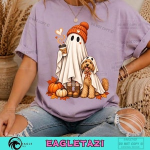 Puede incluir: Una camiseta de color morado claro con un gráfico de un fantasma con gorro naranja que sostiene una taza de café, junto a un cachorro de golden retriever disfrazado de fantasma. Dos calabazas naranjas descansan sobre hojas de otoño.