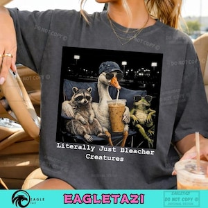 May include: Dark gray t-shirt featuring a graphic of a raccoon, goose, and frog wearing sunglasses and drinking iced coffee. The text "Literally Just Bleacher Creatures" is below the image. The shirt has a relaxed fit.