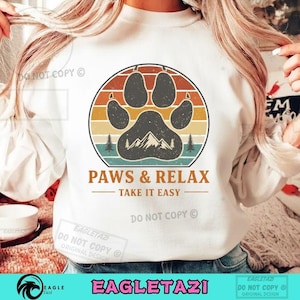 以下が含まれることがあります： レトロな配色で足跡、山、木を描いた円形グラフィックが特徴の白いスウェットシャツ。「PAWS & RELAX - TAKE IT EASY」の文字が下にあります。ブランド名「EAGLETAZI」が一番下にあります。