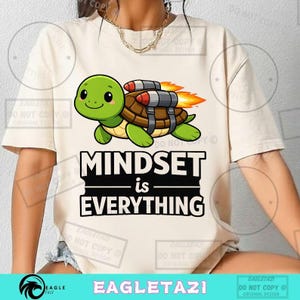Può includere: T-shirt color crema con una tartaruga dei cartoni animati con un jetpack e fiamme, con la scritta "MINDSET is EVERYTHING". La tartaruga è verde e marrone. La maglietta ha maniche corte.