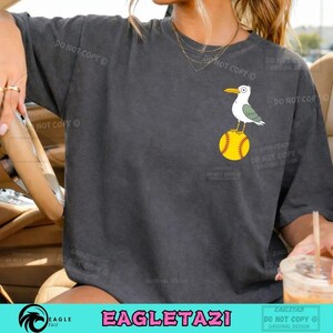 Puede incluir: Camiseta gris oscuro con una gaviota de dibujos animados posada sobre una pelota de softbol amarilla. La gaviota tiene detalles blancos, verdes y amarillos. La camiseta tiene el texto "EAGLETAZI" en la parte inferior.