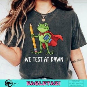 May include: Dark grey t-shirt with a cartoon frog superhero holding a pencil and a notebook. The frog wears a red cape and a red necktie. The text "WE TEST AT DAWN" is printed below the frog.