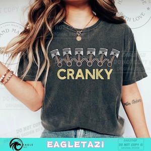 Könnte beinhalten: Dunkelgraues T-Shirt mit einer Grafik von Motor-Kolben über dem Wort "CRANKY" in Gelb. Das T-Shirt hat einen Vintage-Look. Das Design ist mittig auf der Vorderseite des T-Shirts platziert.