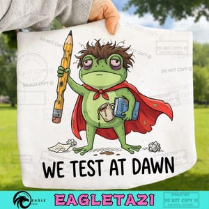 Może przedstawiać: Biała tkanina z kreskówkową żabą w czerwonej pelerynie, trzymającą ołówek i książki. Żaba ma plamę po kawie. Tekst na dole brzmi "WE TEST AT DAWN". Projekt zawiera tekst "EAGLETAZI".