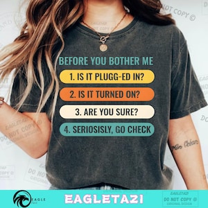 Könnte beinhalten: Dunkelgraues T-Shirt mit dem Text „BEFORE YOU BOTHER ME“ und einer Checkliste zur Fehlerbehebung: „1. IS IT PLUGG-ED IN?“, „2. IS IT TURNED ON?“, „3. ARE YOU SURE?“ und „4. SERIOSISLY, GO CHECK“. Der Text ist farbig.