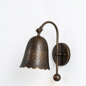 May include: A bronze-colored wall sconce with a bell-shaped shade featuring small perforations. The shade has a scalloped edge and is attached to a curved arm. The sconce includes a round backplate and a decorative ball at the bottom.
