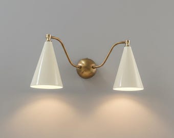 May include: A double wall sconce featuring two off-white conical shades attached to curved brass arms extending from a central circular brass wall mount. The fixture emits a warm downward glow from both light sources, creating a balanced illumination.