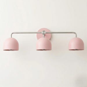 May include: A three-light wall fixture featuring a horizontal brushed nickel bar and a circular wall mount. Three matte pink dome-shaped shades are attached to the bar, each pointing downward to provide focused illumination for interior spaces.