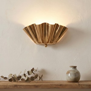 May include: A gold-coloured wall sconce with a shell-like design. The light fitting is mounted on a white wall and features a small, spherical detail at the base. A wooden shelf below holds a small vase and dried foliage.